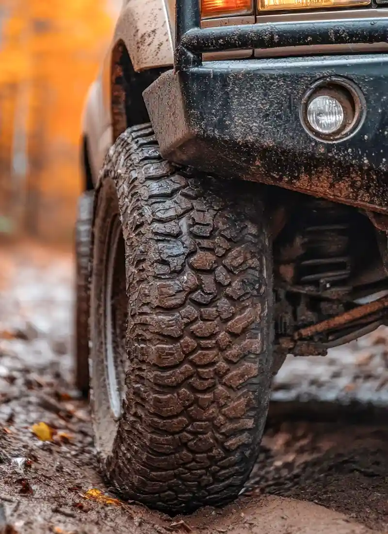 Muddy all-terrain tire on off-road 4x4 truck, autumn trail; rugged tread, fog light, suspension - Automotive Expressions