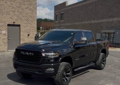 Automotive Expressions black RAM 1500 crew-cab with custom wheels, all-terrain tires, blacked-out grille near brick warehouse