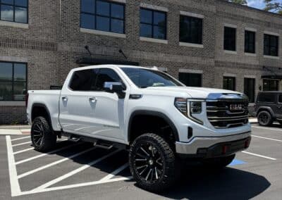 Automotive Expressions lifted white GMC Sierra on black custom off-road wheels, window tint, red tow hooks at brick office